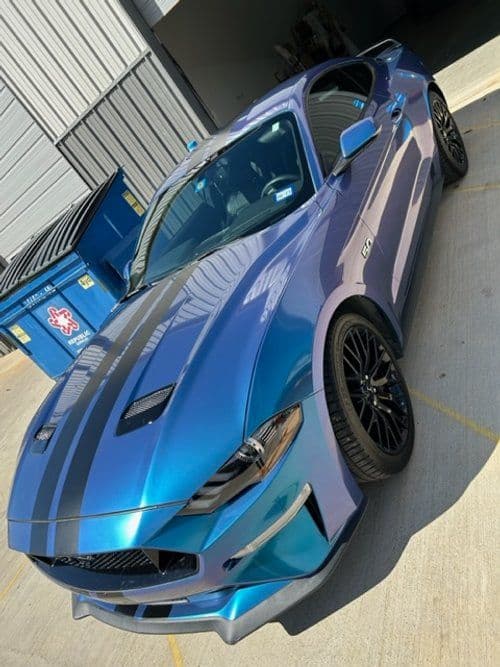 A blue mustang is parked in a parking lot next to a dumpster